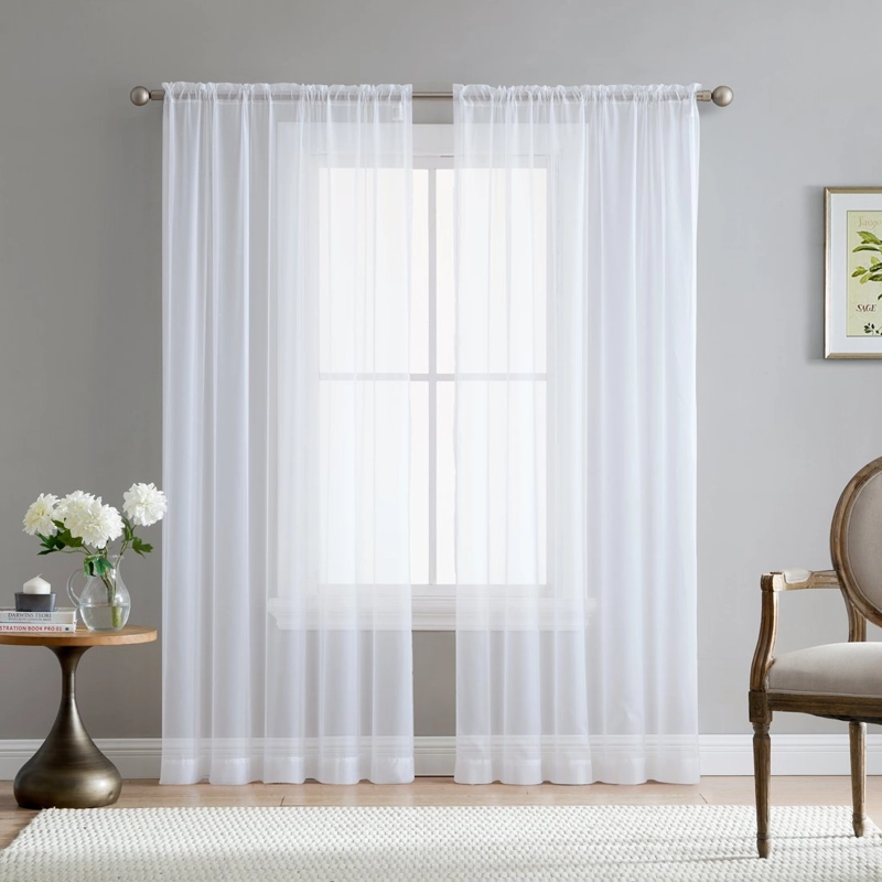 Sheer white curtains hang over a bright window in a room with a small table, flowers