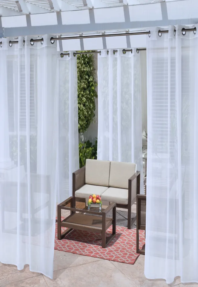 White sheer curtains on a pergola frame an outdoor seating area with a sofa, coffee table, and red rug.
