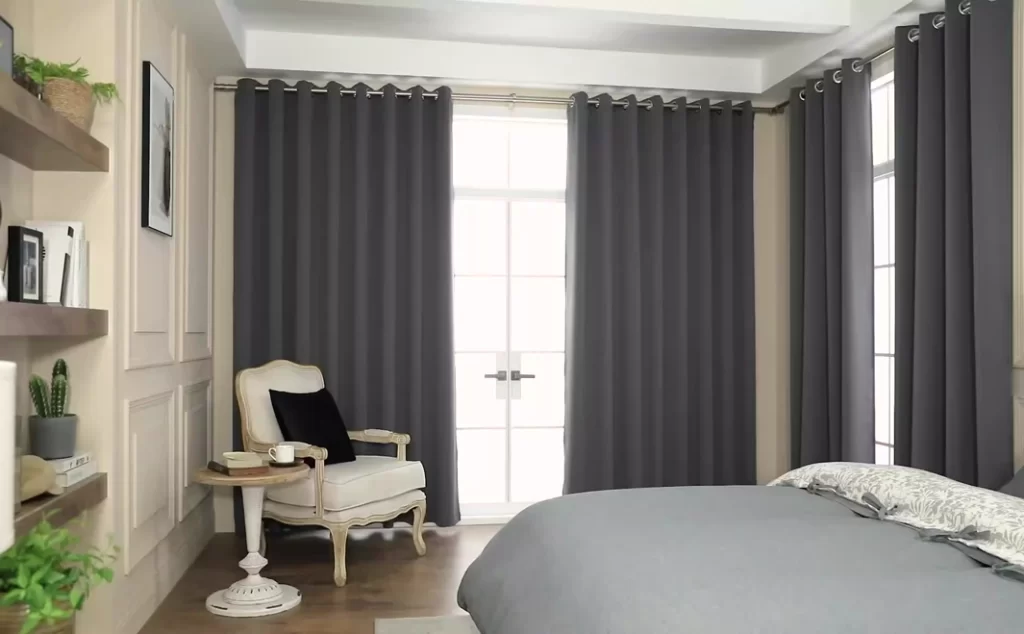 Dark grey blackout curtains in a modern bedroom with a cream armchair and light grey bedding.