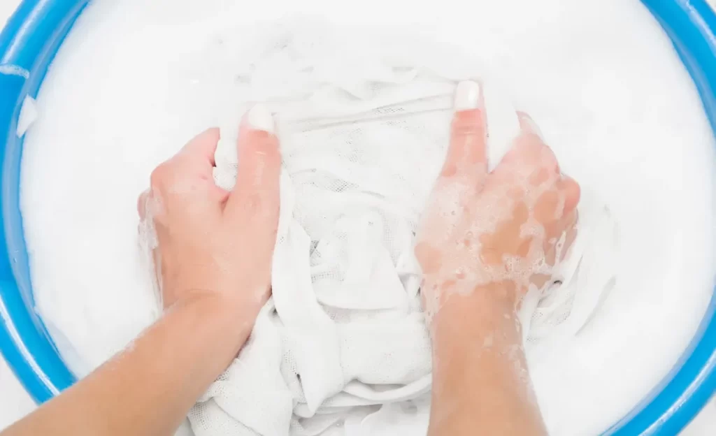 Hands hand-washing a white garment in a blue basin with suds.