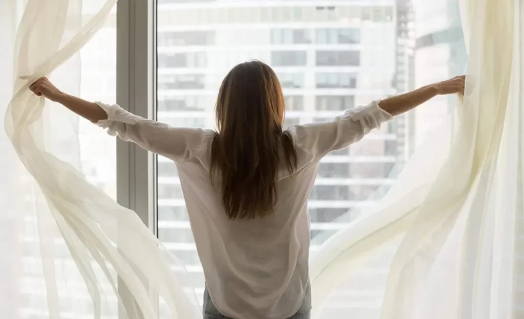 A person from behind pulls back sheer white curtains to reveal a cityscape.