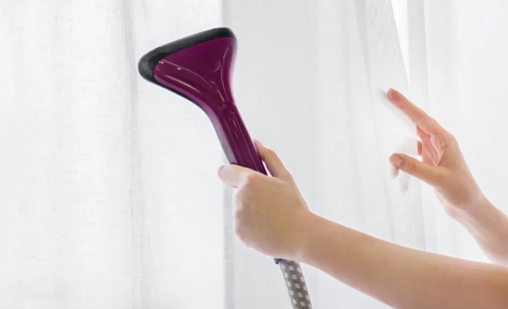 A person steams a white sheer curtain with a purple handheld steamer.